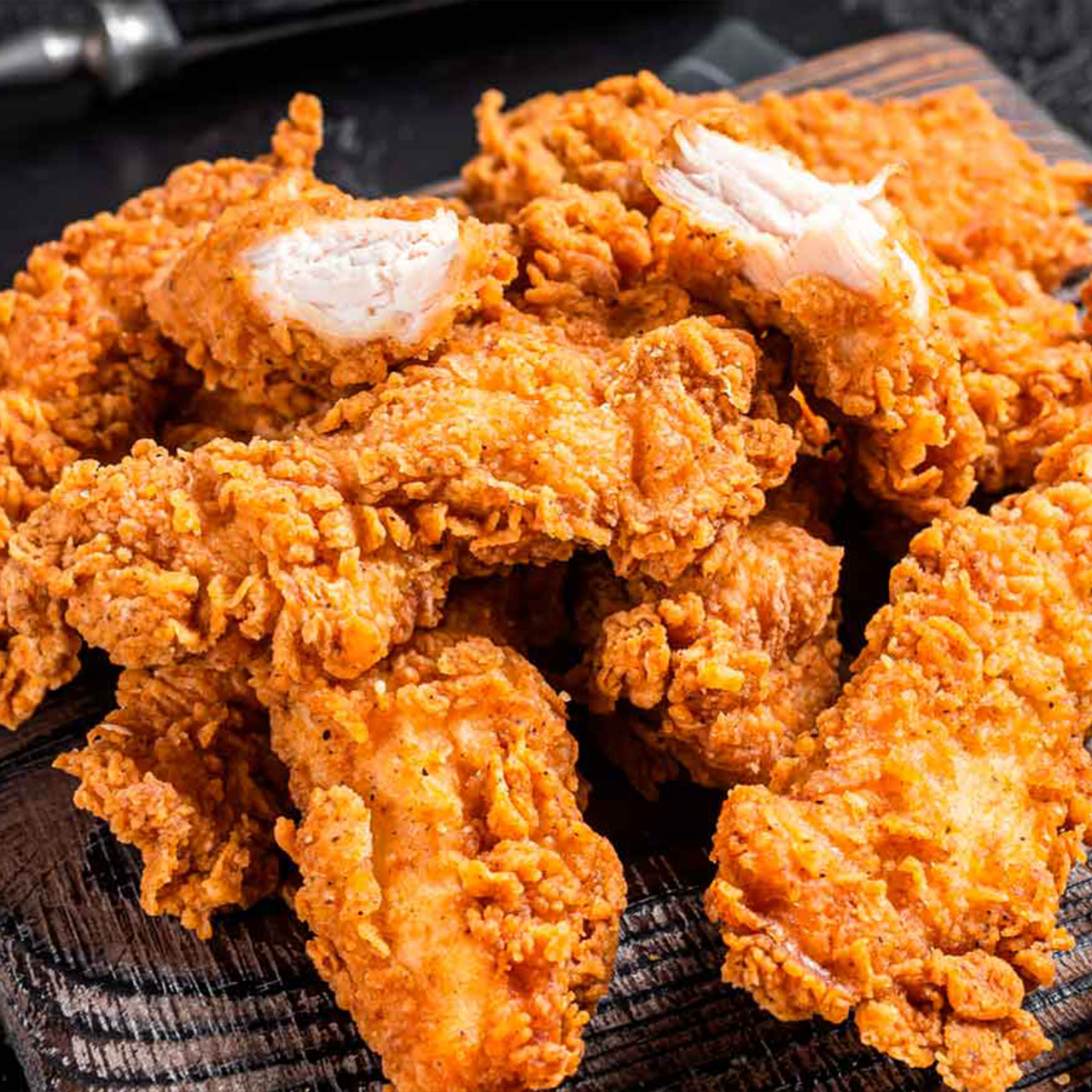 Ein Haufen goldbrauner, knuspriger Hähnchen-Tender auf einem dunklen Holzbrett. Einige Stücke sind aufgebrochen und zeigen das weiße, gekochte Fleisch.